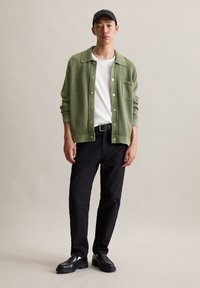 Green knit cardigan with button closure over a white t-shirt, paired with black pants and black shoes. Model wears a black cap.