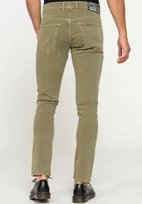 Jeans slim fit vert olive avec cinq poches, coutures visibles et patch en cuir sur la ceinture, associés à des chaussures noires.