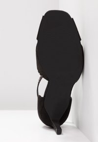 Sandale à talon en daim noir avec bout carré, bride cheville cloutée et talon fin, vue du côté de la semelle, reposant contre une surface blanche.