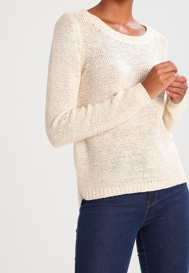 Personne portant un pull en maille crème à manches longues et un jean bleu foncé, ajustant une manche avec les deux mains.