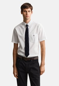 Young man wearing a white short-sleeve button-up shirt, dark blue polka dot tie, patterned belt, and black pants, standing against white background.