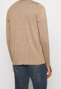 Beige neulepusero pyöreällä pääntiellä ja resorihihansuuilla. Siinä on teksturoitu viimeistely ja pieni logoyksityiskohta takana.