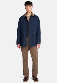 Mörkblå jacka med en brun krage, med två fickor framtill, lätt struktur och knäppning med knappar. Kombinerad med beige byxor och bruna skor.