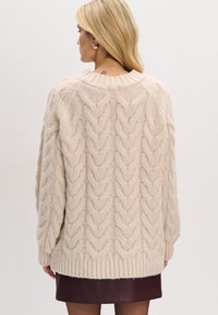 Beżowy sweter w sploty z luźnym krojem, fakturowanym wzorem, prążkowanym dolnym brzegiem i opuszczonymi ramionami, noszony na bordowej skórzanej spódnicy.