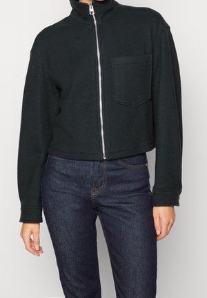 Black cropped zip-up jacket with chest pocket worn with dark blue jeans on a person against a light background.
