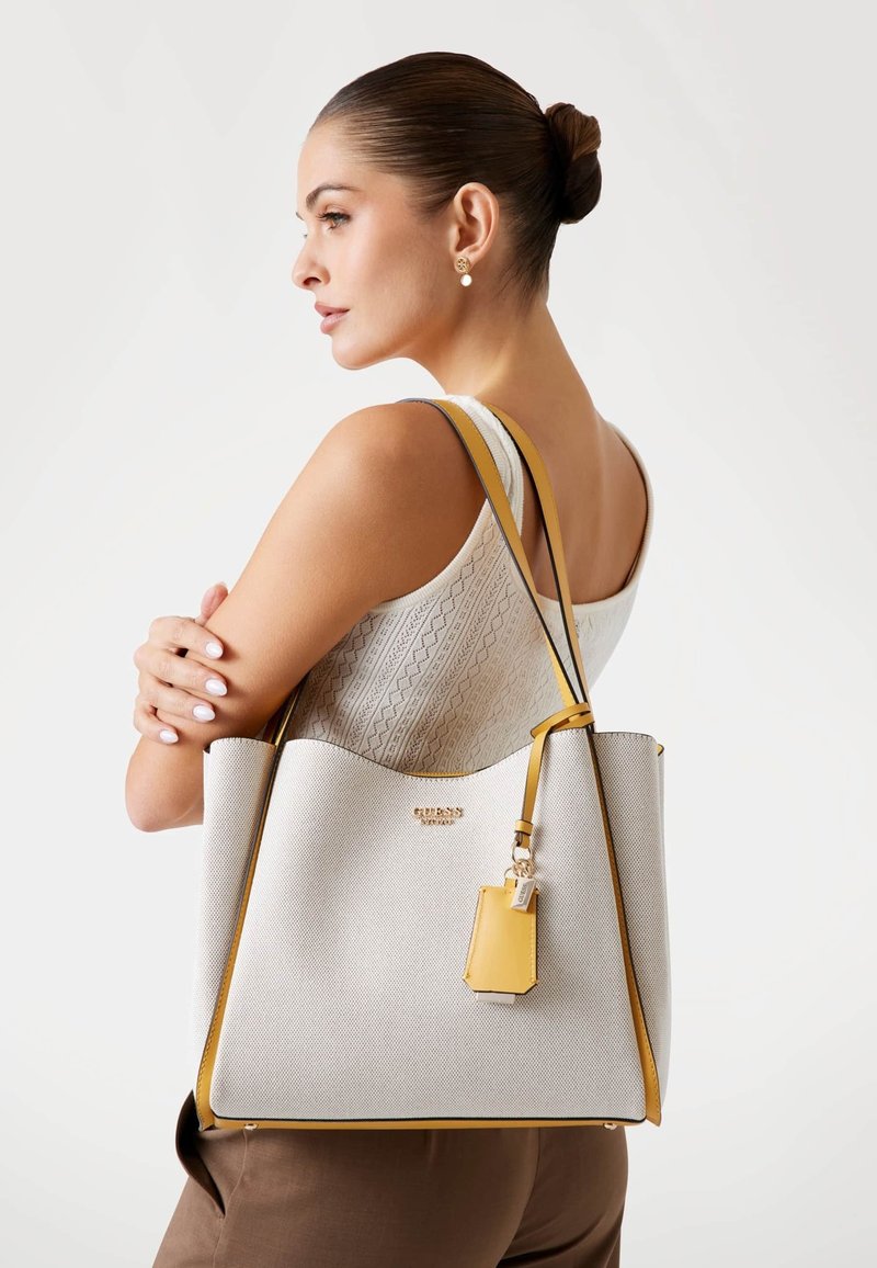 Grand sac fourre-tout gris clair avec des accents jaunes et une finition texturée. Présente un logo doré et un petit charme détachable.