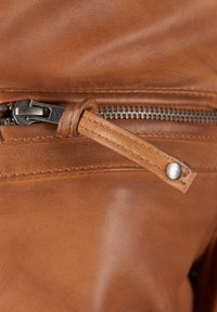 Detail van een bruine leren tas met een rits met een zilveren trekker, een gestructureerd oppervlak en een contrasterende schouderband met een drukknoopaccent.