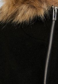 Sort uldjakke med en tan pelskrave og en sølv lynlås. Har en glat tekstur og et tætsiddende design.