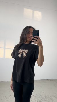Melna kokvilnas T-kreklu ar leoparda raksta loku grafiku priekšā. Valkāts ar melnajām legingām labi apgaismotā iekštelpās.