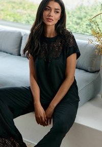 Hosszú sötét hajú nő fekete csipkefelsőt és nadrágot visel, fehér párkányon ül, szürke csíkos kanapé mellett, az ablak közelében.