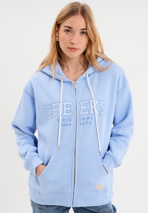 Kobieta nosząca jasnoniebieską bluzę z kapturem na zamek z napisem "CREEKS ORIGINAL since 1974", ręce w kieszeniach i niebieskie dżinsy na białym tle.