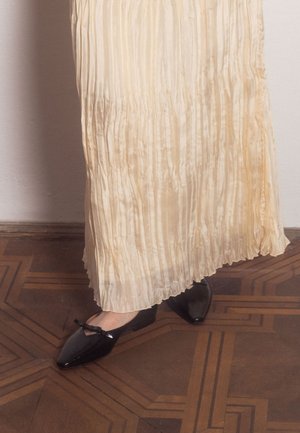 Jupe plissée couleur crème portée avec des ballerines noires brillantes à bouts pointus ornées de petits nœuds, sur un parquet en bois.