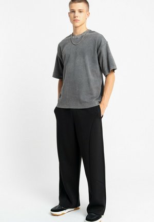 Młody mężczyzna stoi, ubrany w luźną szarą koszulę z krótkim rękawem, czarne szerokie spodnie, czarne buty z białą podeszwą oraz srebrny naszyjnik z łańcuszkiem.