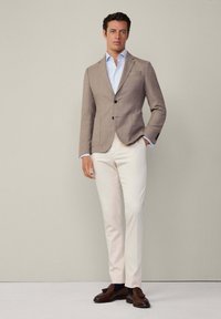 Blazer de texture marron, chemise bleu clair, pantalon crème et mocassins marron. Le blazer présente deux boutons et une poche unique.