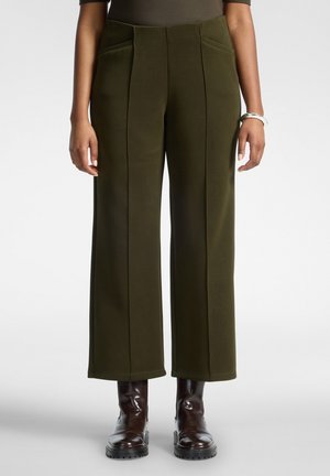 Femme portant un pantalon large vert olive avec des coutures frontales et des bottines marron, debout les bras détendus le long du corps.