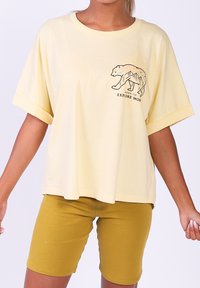 Gelbes Baumwoll-T-Shirt mit kurzen Ärmeln, das ein Bär-Grafik und den Text "Born to Explore More" auf der Vorderseite zeigt. Kombiniert mit senfgelben Shorts.