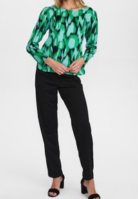 Blouse verte à motifs avec des manches longues, des accents noirs et un décolleté carré, associée à un pantalon noir et des sandales à talons hauts noires.