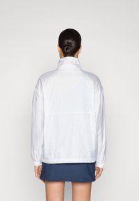 Hvid vindjakke med høj krave, elastiske manchetter og en afslappet pasform. Subtil tekstur og mønster synligt på stoffet.