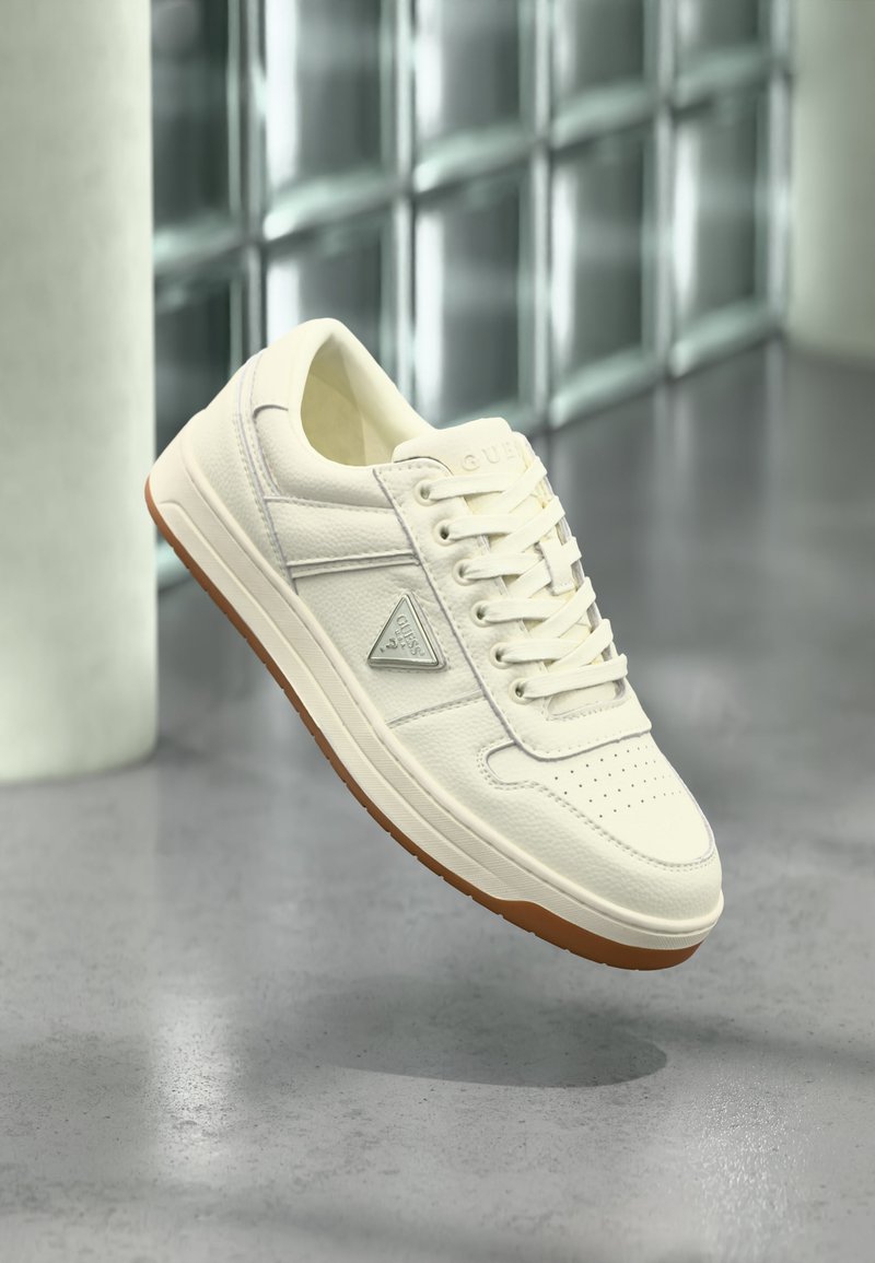 Λευκό sneaker χαμηλού προφίλ με καουτσούκ σόλα, μπροστινό δέσιμο με κορδόνια και τριγωνικό λογότυπο Guess στο πλάι, παρουσιάζεται με φόντο θολό από γυάλινους κύβους.