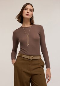 Bruine lange mouwen geribbelde top gecombineerd met hooggetailleerde, op maat gemaakte bruine broeken, met een gouden ronde riemgesp en een gelaagde gouden ketting.