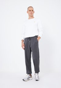 Graue Jogginghosen mit Taschen und einem elastischen Bund, kombiniert mit einem weißen Langarmshirt mit einem gelben Logo. Sportliche Schuhe runden das Outfit ab.