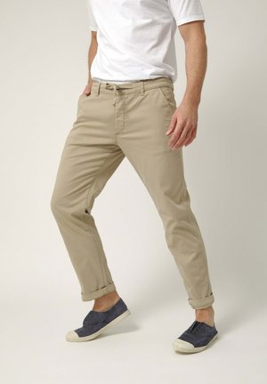Hombre con pantalones beige con puños, camiseta blanca y zapatillas slip-on azul marino, de pie con las piernas separadas sobre un fondo liso.