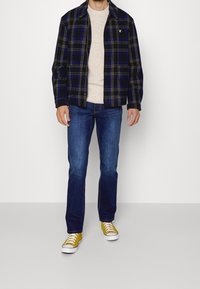 Chemise à carreaux bleu marine et jaune, léger pull, jean bleu et baskets jaunes. La chemise a un col et des poches avant.