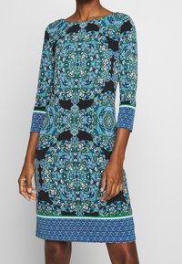 Långärmad klänning med svart bakgrund som har intrikata blå och gröna blommönster, texturerad tyg och rak passform.