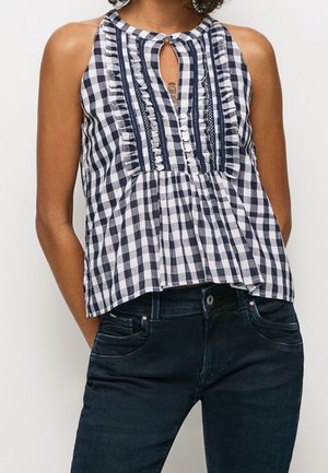 Kvinde iført ærmeløs marineblå og hvid ternet bluse med flæser og knapper, kombineret med mørkeblå jeans.