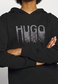 Czarna bluza z kapturem z literami z cyrkonii piszącymi "HUGO" w gradientowym efekcie. Długie rękawy, luźny krój, średnia grubość materiału.