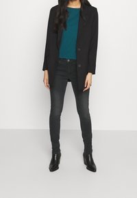 Blazer negro sobre una blusa azul verdosa, combinado con jeans ajustados oscuros y botas de tacón negras por el tobillo. Texturas suaves y un diseño ajustado en todo.