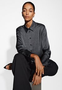 Camicia nera, in tessuto strutturato, con colletto, chiusura a bottoni e maniche lunghe. Modello in pantaloni neri, seduto, con le mani appoggiate sulle ginocchia.