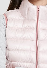 Hellrosa, gesteppte Weste mit hohem Kragen und Reißverschluss; verfügt über eine glänzende Textur und ein gestepptes Muster; getragen über einem grauen Oberteil.