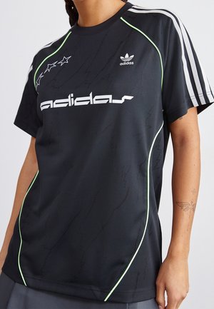 Czarny t-shirt Adidas z białym logo, trzema białymi paskami na rękawach, neonowo zielonym przeszyciem oraz wzorami gwiazd w okolicy ramienia, noszony przez osobę.