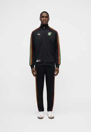 Man die staat en een zwart trainingspak draagt met rode, gele en groene strepen op de mouwen en broek, witte sneakers en logo's op het jack en de broek.
