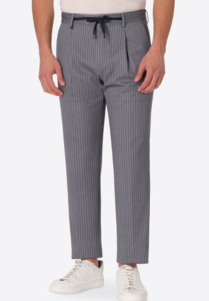 Uomo che indossa pantaloni grigi a righe sottili con coulisse nera e sneakers bianche, in piedi davanti a uno sfondo semplice.