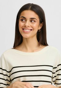 Suéter crema con rayas negras horizontales. Presenta un escote amplio y una textura suave. El modelo muestra una leve sonrisa y aretes de aro.