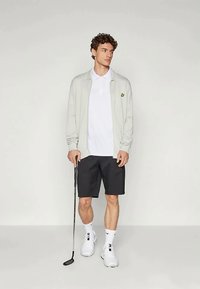 Cardigan lavorato a maglia grigio chiaro sopra una polo bianca, abbinato a pantaloni corti neri. In piedi su uno sfondo neutro, con in mano una mazza da golf.