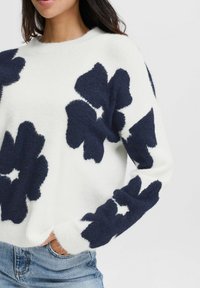 Pullover, weiß mit großen marineblauen Blumenmustern, weicher Textur, Rundhalsausschnitt, langen Ärmeln, gerippten Bündchen und Saum, getragen zu blauen Jeans.