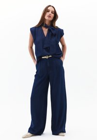 Námořnická modrá blůza s uvázanou mašlí u výstřihu, v kombinaci s širokými námořnickými džínovými kalhotami a světlými, texturovanými botami.