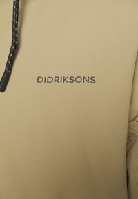 Gros plan sur un tissu beige avec le logo DIDRIKSONS noir et une partie d’un cordon noir et blanc.