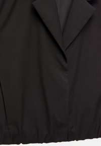 Veste noire cintrée avec col à revers cranté, fermeture par boutons et un ourlet légèrement froncé en bas.