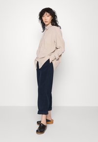 Camicia beige con bottoni, pantaloni in corduroy neri e zoccoli in legno neri. Vestibilità ampia con orli arrotolati e dettagli sulla tasca frontale.