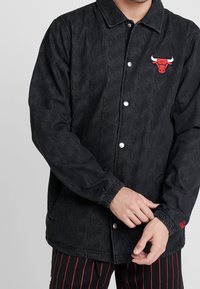 Person som justerar ärmslutet på en svart Chicago Bulls-jacka med upprepande logotext och röd tjur-emblem, ihop med svarta shorts med röda ränder.