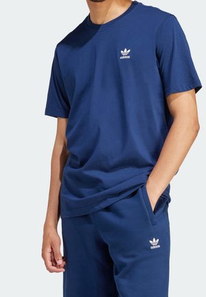 Mies, jolla tumma sininen Adidas t-paita ja housut valkoisilla Adidas-logoilla, toinen käsi taskussa ja toinen rentona sivulla.