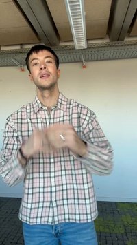 Joven con cabello oscuro y corto que lleva una camisa de cuadros rosa y verde y vaqueros azules, haciendo gestos con las manos desenfocadas en interiores.