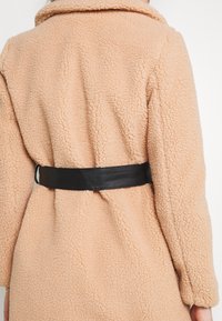 Faux fur frakke i lys fersken med en struktureret, fluffy overflade. Har et sort, glat læderbælte i taljen, der skaber et indsnævret look.