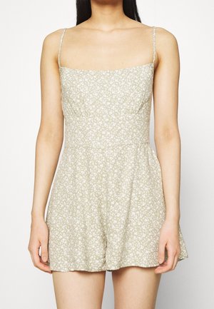 Romper w kwiatowy wzór w jasnym beżu z cienkimi ramiączkami, kwadratowym dekoltem, ewidentnie taliowanym pasem i luźnymi szortami. Materiał wydaje się być miękki i teksturowany.