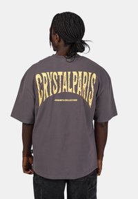 Šedé oversized tričko s velkým žlutým nápisem "CRYSTALPARIS" na zadní straně, pod kterým je "CHAMPS COLLECTION". Krátké rukávy, bavlněný materiál.