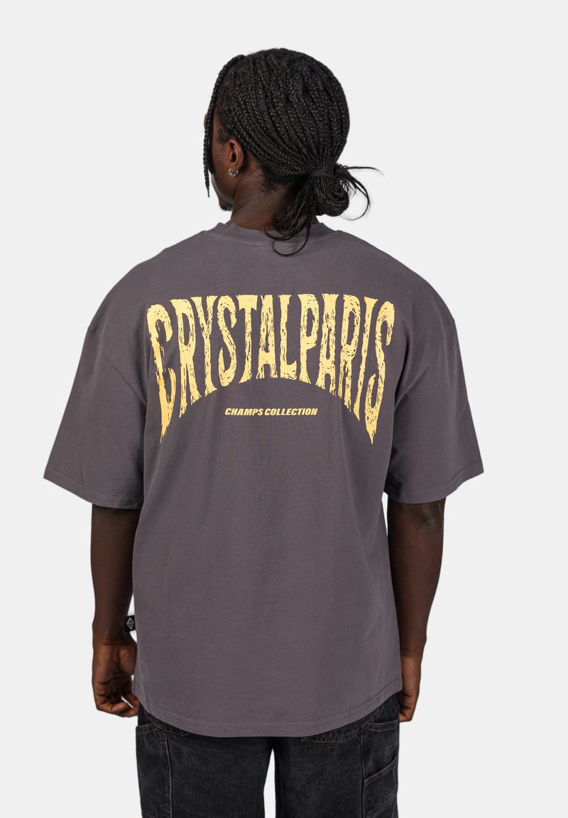 Šedé oversized tričko s velkým žlutým nápisem "CRYSTALPARIS" na zadní straně, pod kterým je "CHAMPS COLLECTION". Krátké rukávy, bavlněný materiál.